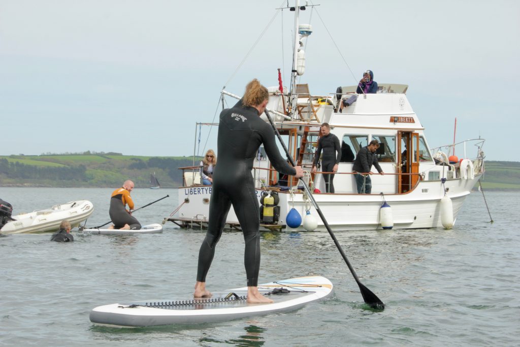 stand up paddle boarder on a boat trip near Mylor, Falmouth, with Liberty Boat Charter in the background.